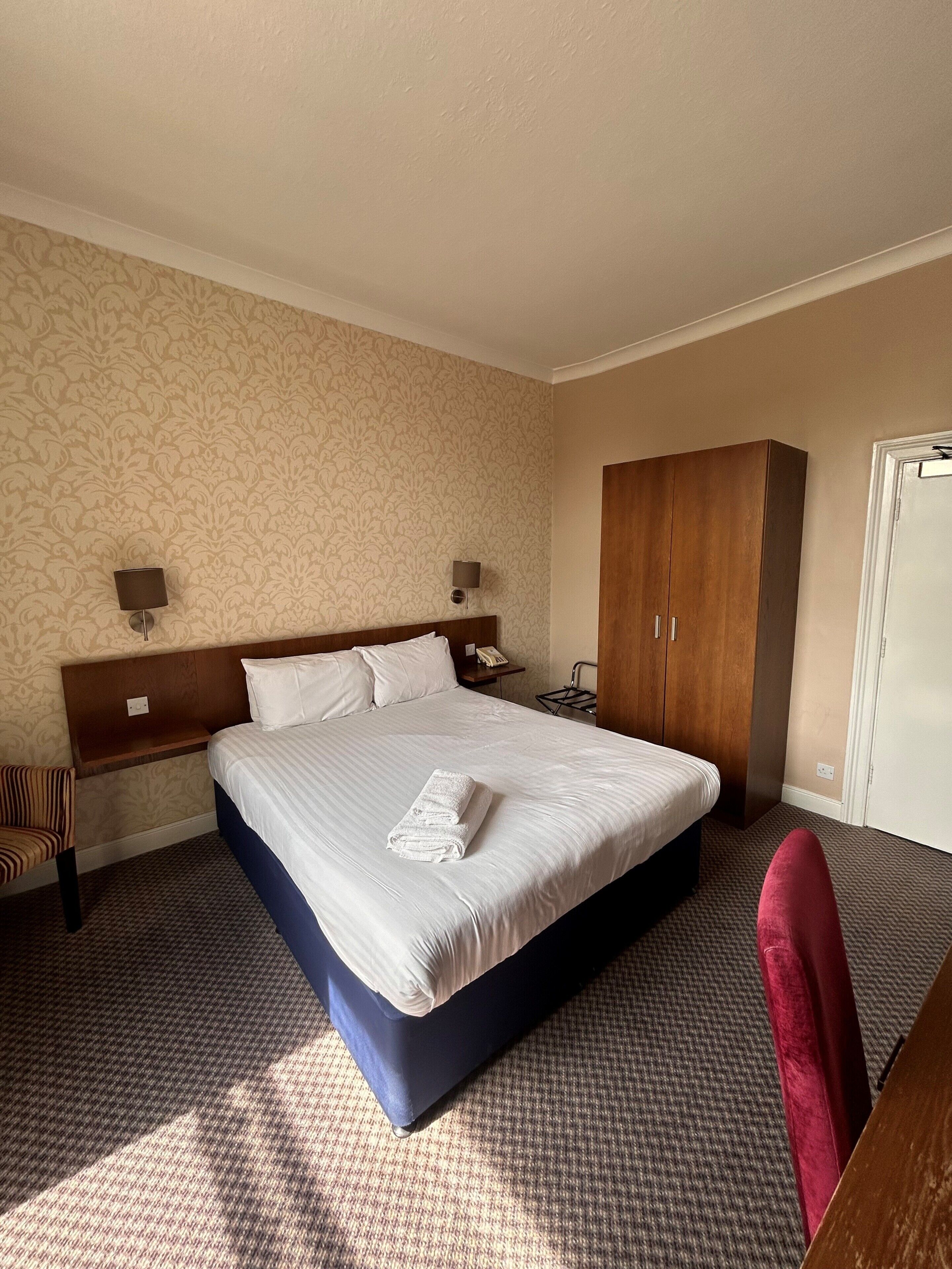 A hotel room with a large bed, a wooden wardrobe, a desk, a chair, and a patterned wallpaper.