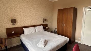 A hotel room with a large bed, a wooden wardrobe, a desk, a chair, and a patterned wallpaper.