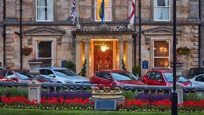 A historic building with a grand entrance, multiple flags, and a parking area with several cars. - Crown Hotel Harrogate (Harrogate)