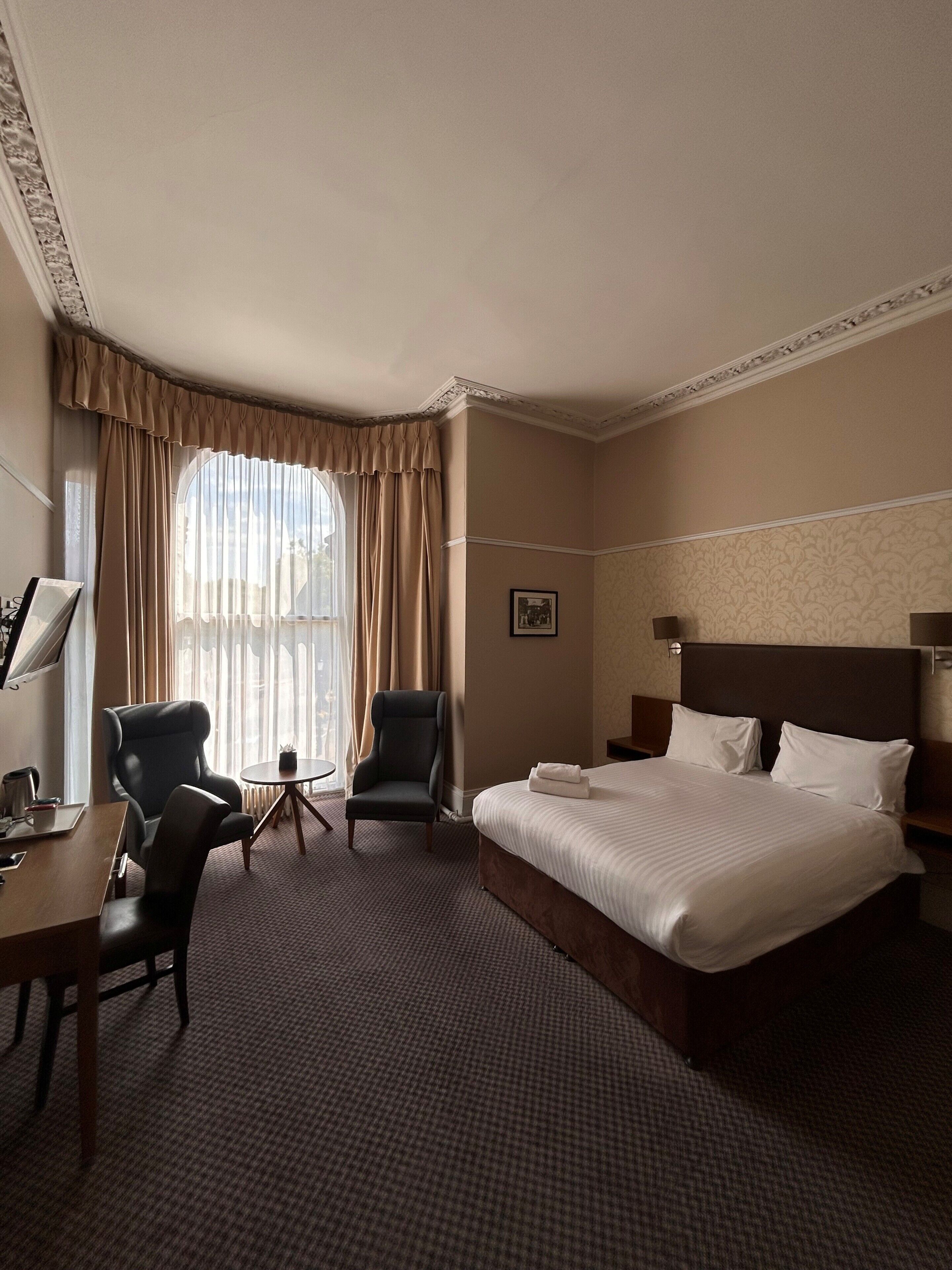 Habitación de hotel con una cama grande, dos sillones, una mesita y un ventanal con cortinas.