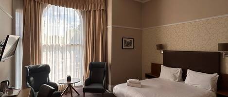 Une chambre d’hôtel avec un grand lit, deux fauteuils, une petite table et une fenêtre avec des rideaux.