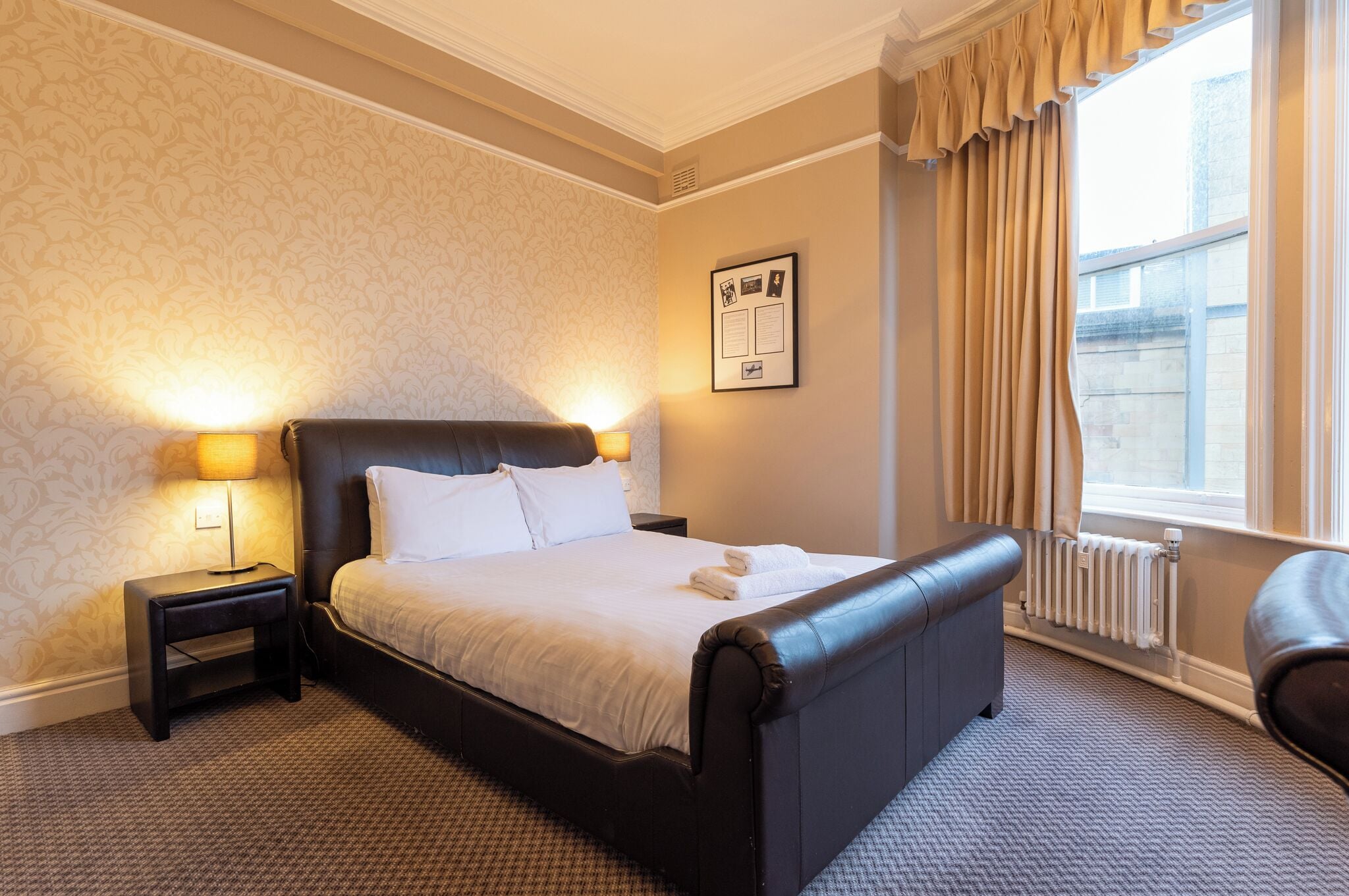 A hotel room with a large bed, two bedside lamps, a radiator, and a window with curtains.