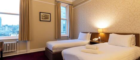 A hotel room with two beds, a radiator, a window with curtains, a framed picture, and a bedside lamp.
