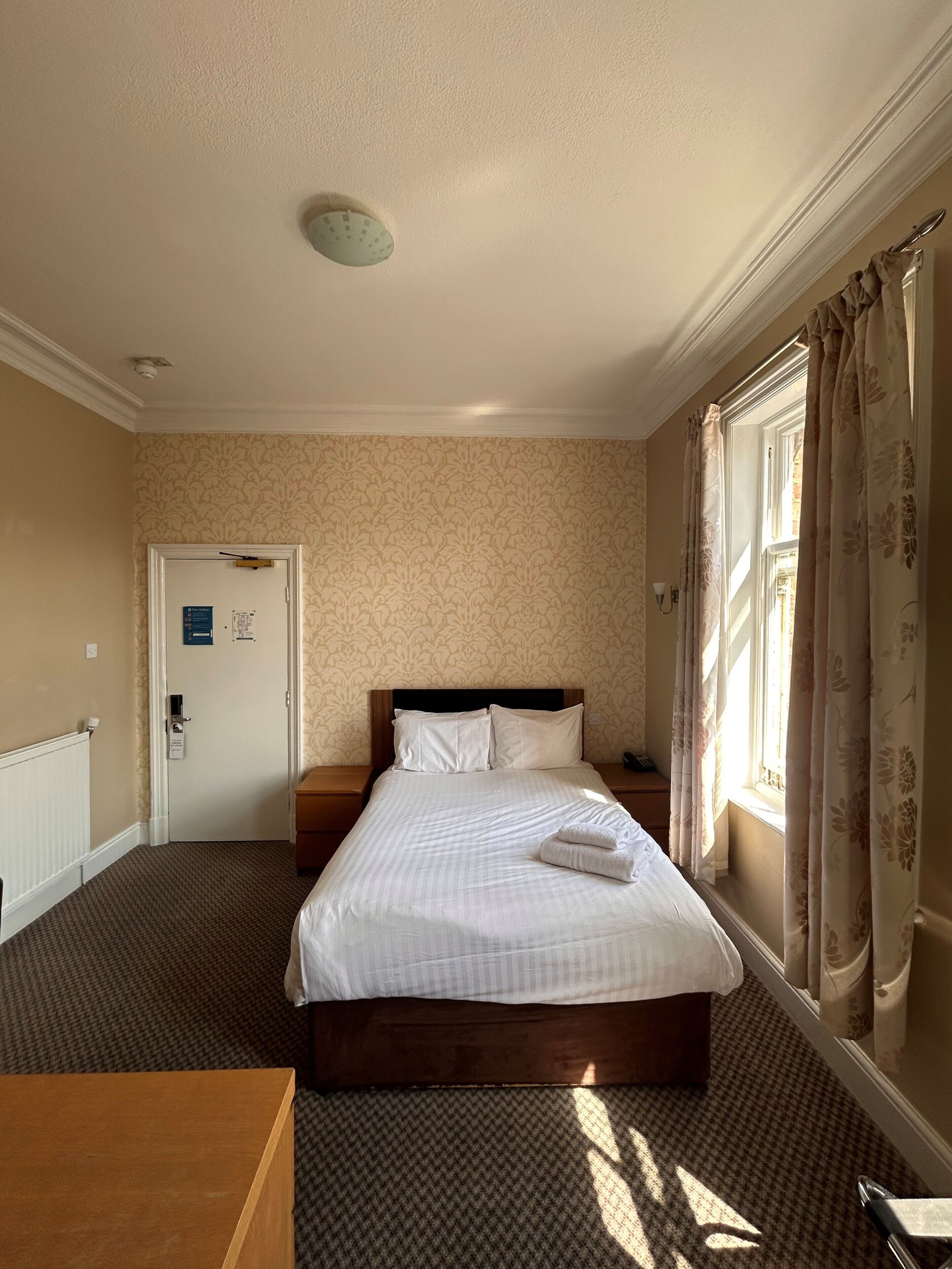 Une chambre d’hôtel avec un lit, une table de chevet en bois, une lampe de chevet, une fenêtre avec des rideaux et une porte avec une pancarte.
