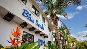 L’extérieur de l’hôtel Blue Sands se distingue par sa façade blanche ornée d’une enseigne bleue, le tout étant entouré d’une végétation tropicale luxuriante et de palmiers.