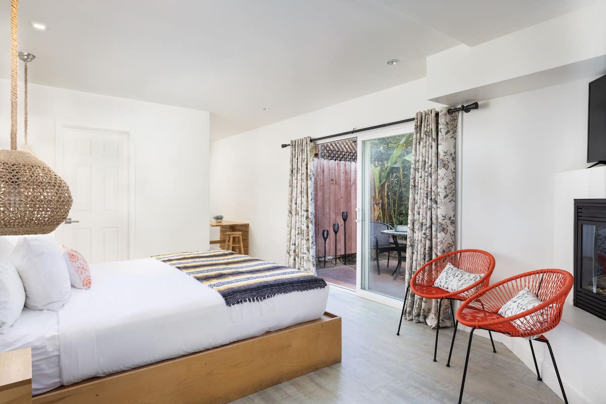A bedroom with a bed, a desk, two red chairs, and a sliding glass door leading to an outdoor area.