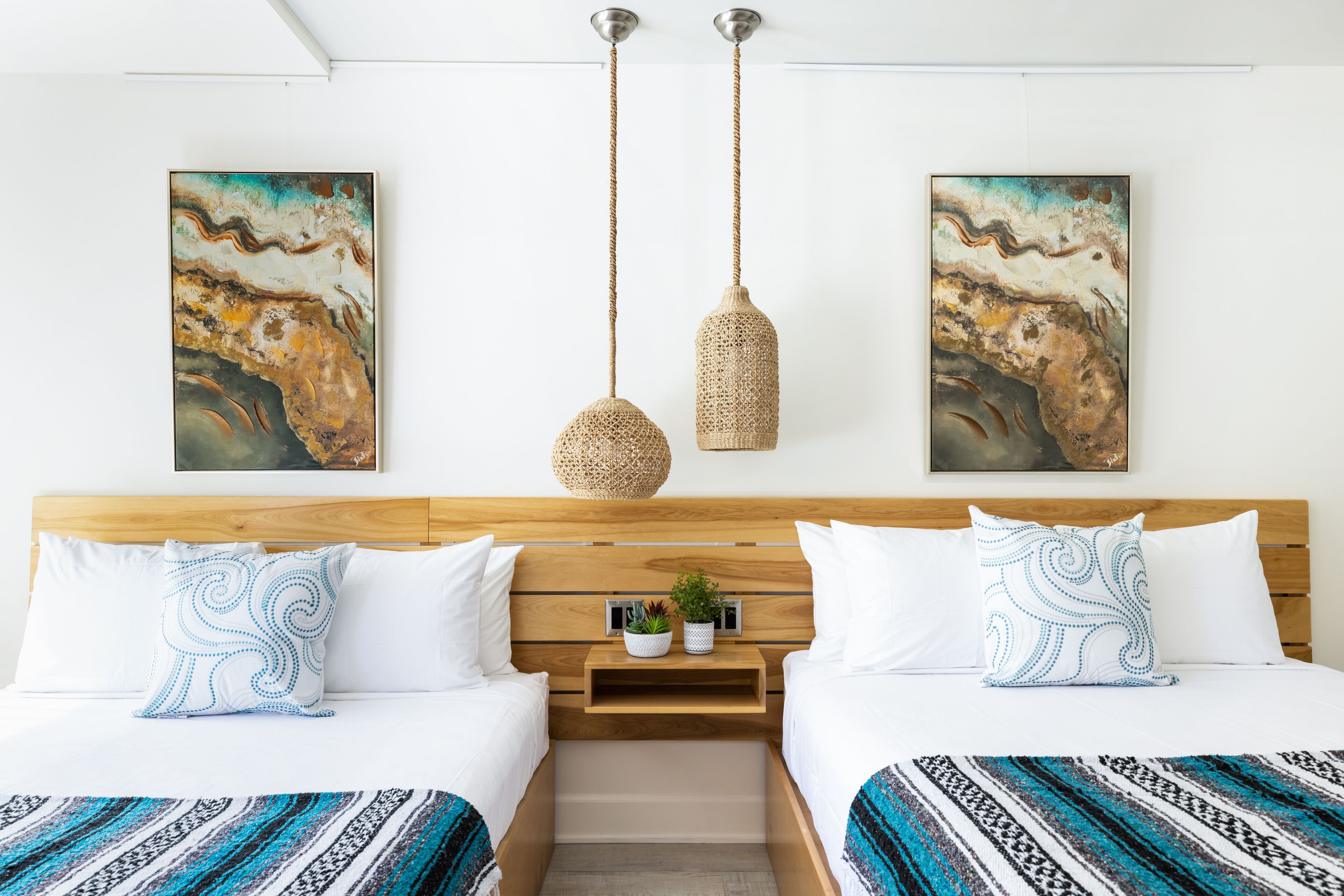A hotel room with two beds, a wooden headboard, a hanging woven light fixture, and two abstract paintings on the wall.