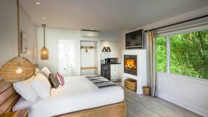 Une chambre moderne avec un grand lit, une cheminée et une vue sur la verdure par la fenêtre.