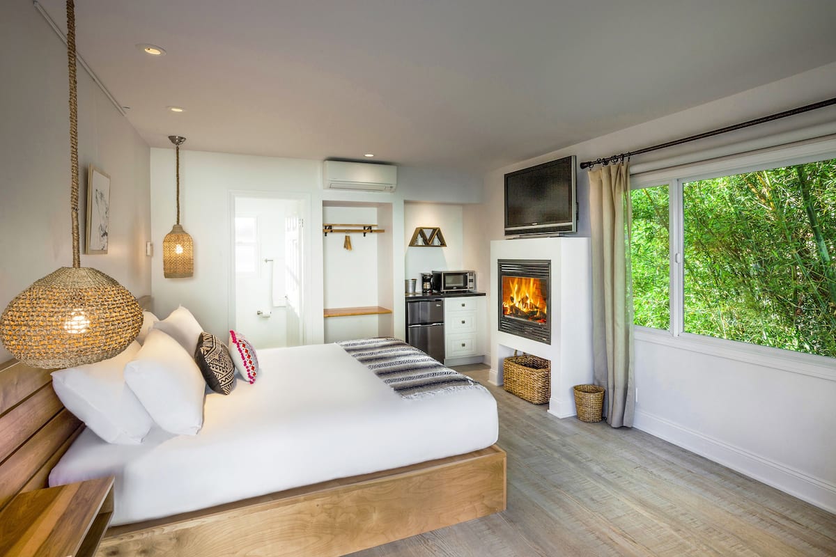 A modern bedroom with a large bed, a fireplace, and a view of greenery through the window.