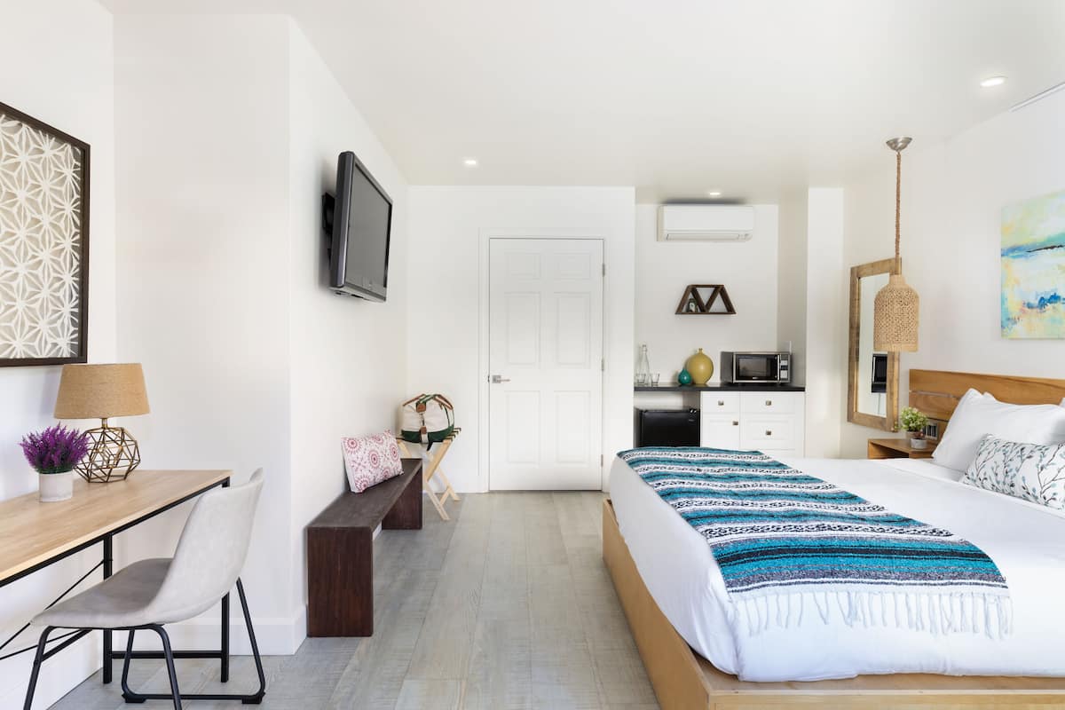 A modern hotel room with a wooden desk, a comfortable chair, a bed with a patterned blanket, a television, a small kitchenette, and a decorative wall piece.