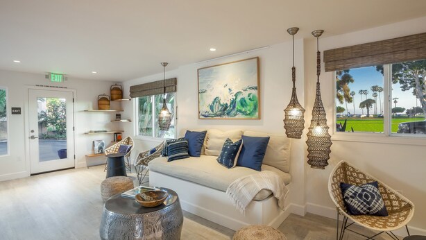 Un salon moderne avec un canapé beige, des coussins bleus, des fauteuils en osier et une grande fenêtre donnant sur des palmiers.