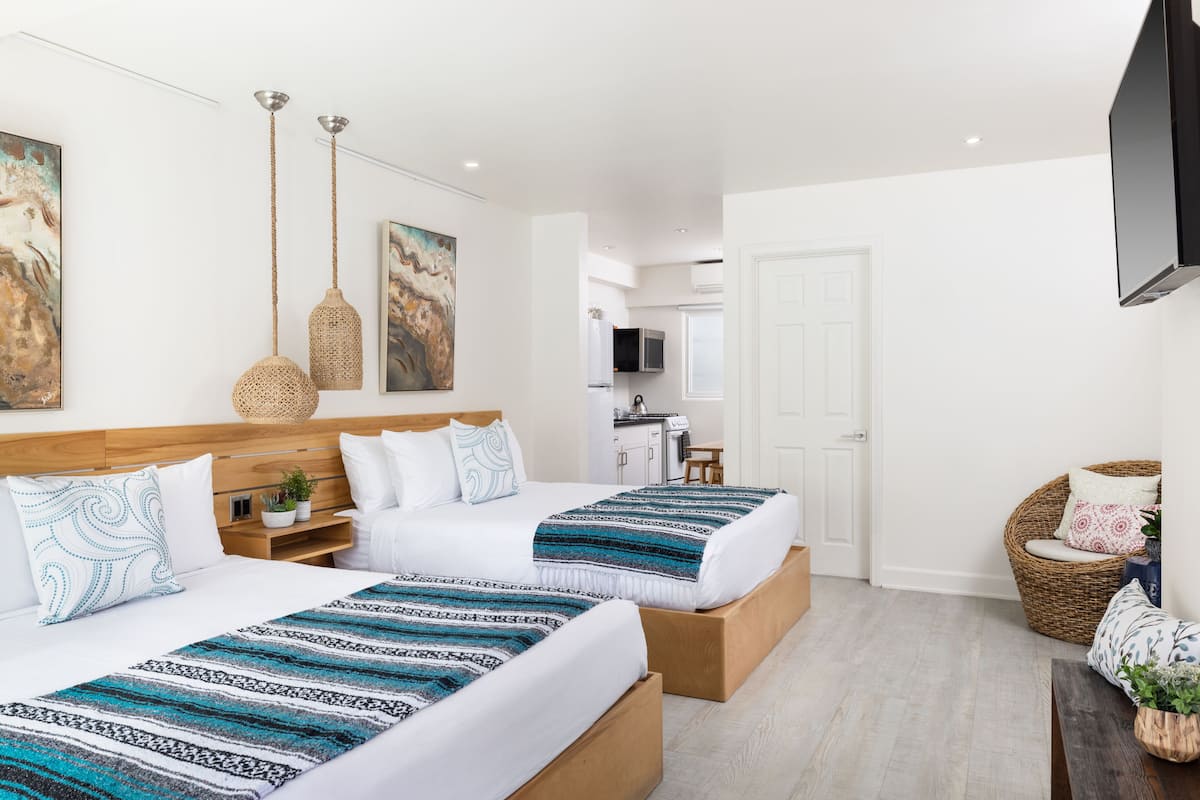 A hotel room with two beds, a wooden headboard, a TV, a kitchen area, and a wicker chair.