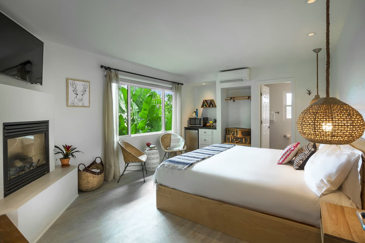 A modern bedroom with a large bed, a fireplace, a dining area, and a view of lush greenery.