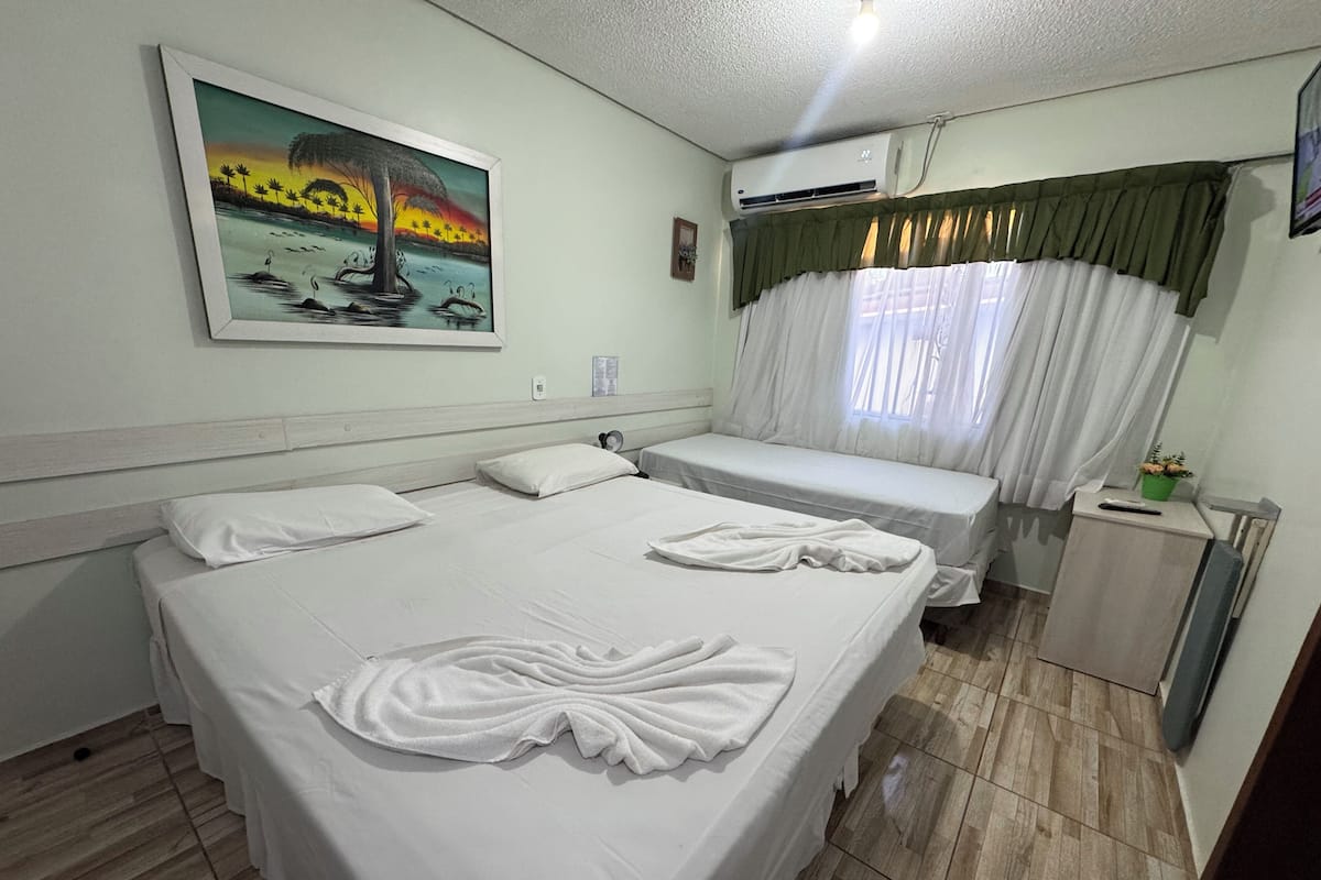 A hotel room with two beds, a painting on the wall, an air conditioner, and a window with curtains.