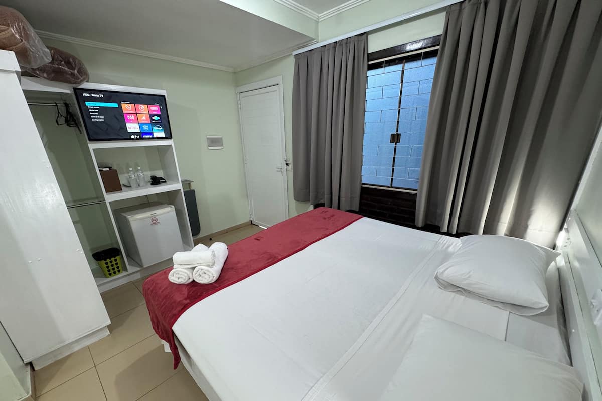 A hotel room with a bed, a TV on a stand, a mini-fridge, a shelf with toiletries, and a window with curtains.