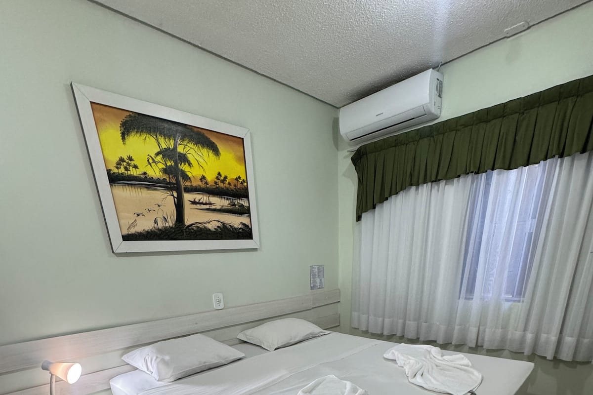 A neatly arranged hotel room with a large bed, a nightstand with a lamp, a painting on the wall, and an air conditioning unit.