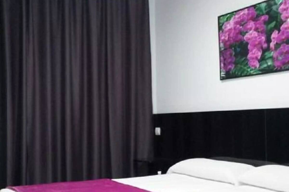 A hotel room with a bed, a large window with curtains, and a wall-mounted painting of flowers.