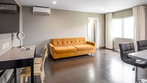 A modern living room with a sofa, a dining table, and chairs. There are air conditioning units on the wall, a mirror on the table, and a window with curtains.