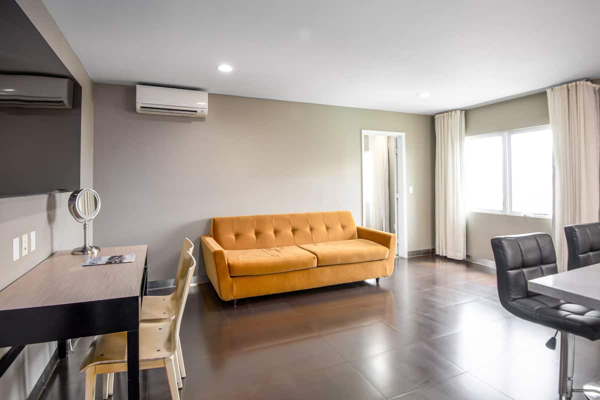 A modern living room with a sofa, a dining table, and chairs. There are air conditioning units on the wall, a mirror on the table, and a window with curtains.
