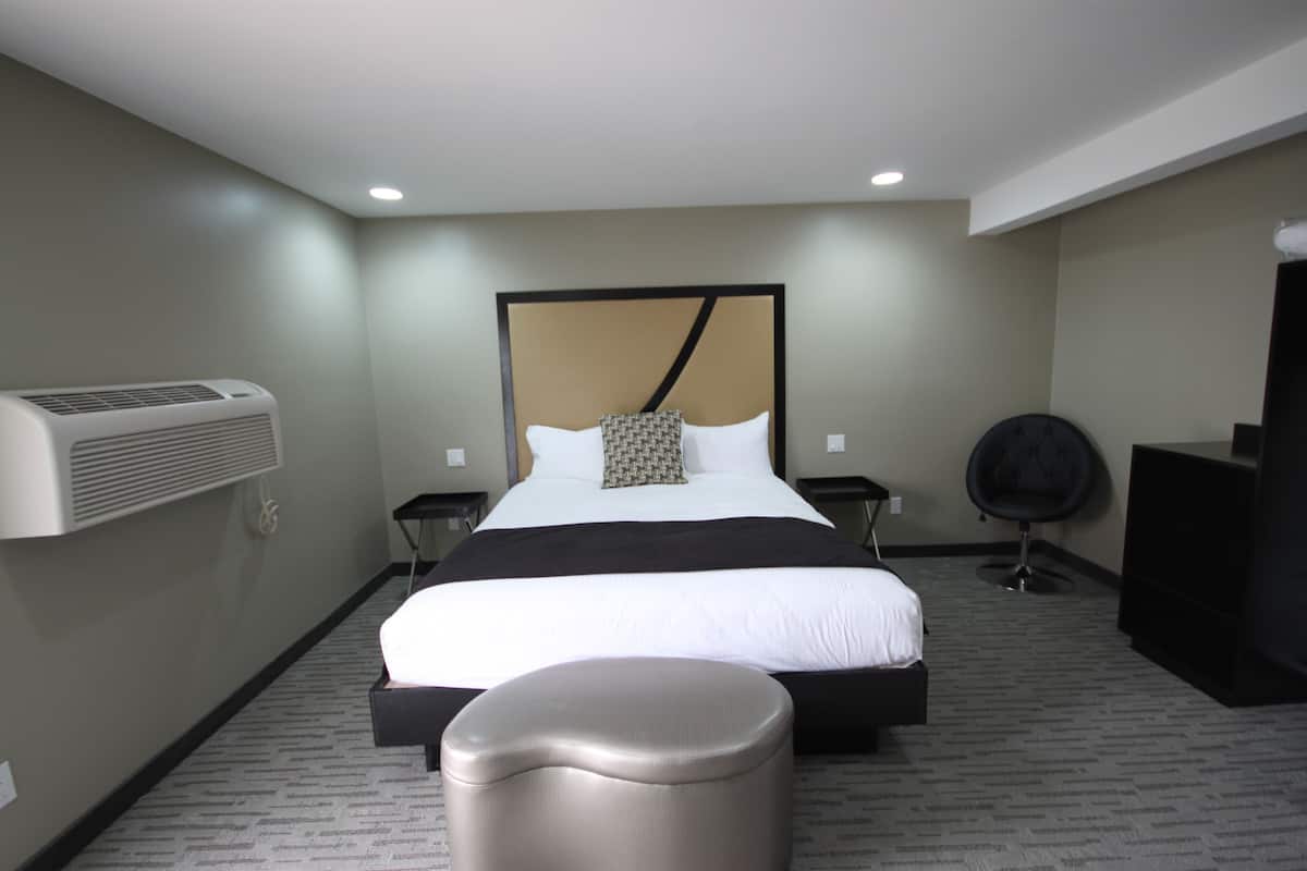 A modern hotel room with a large bed, a bedside table, a chair, an air conditioning unit, and a flat-screen TV.