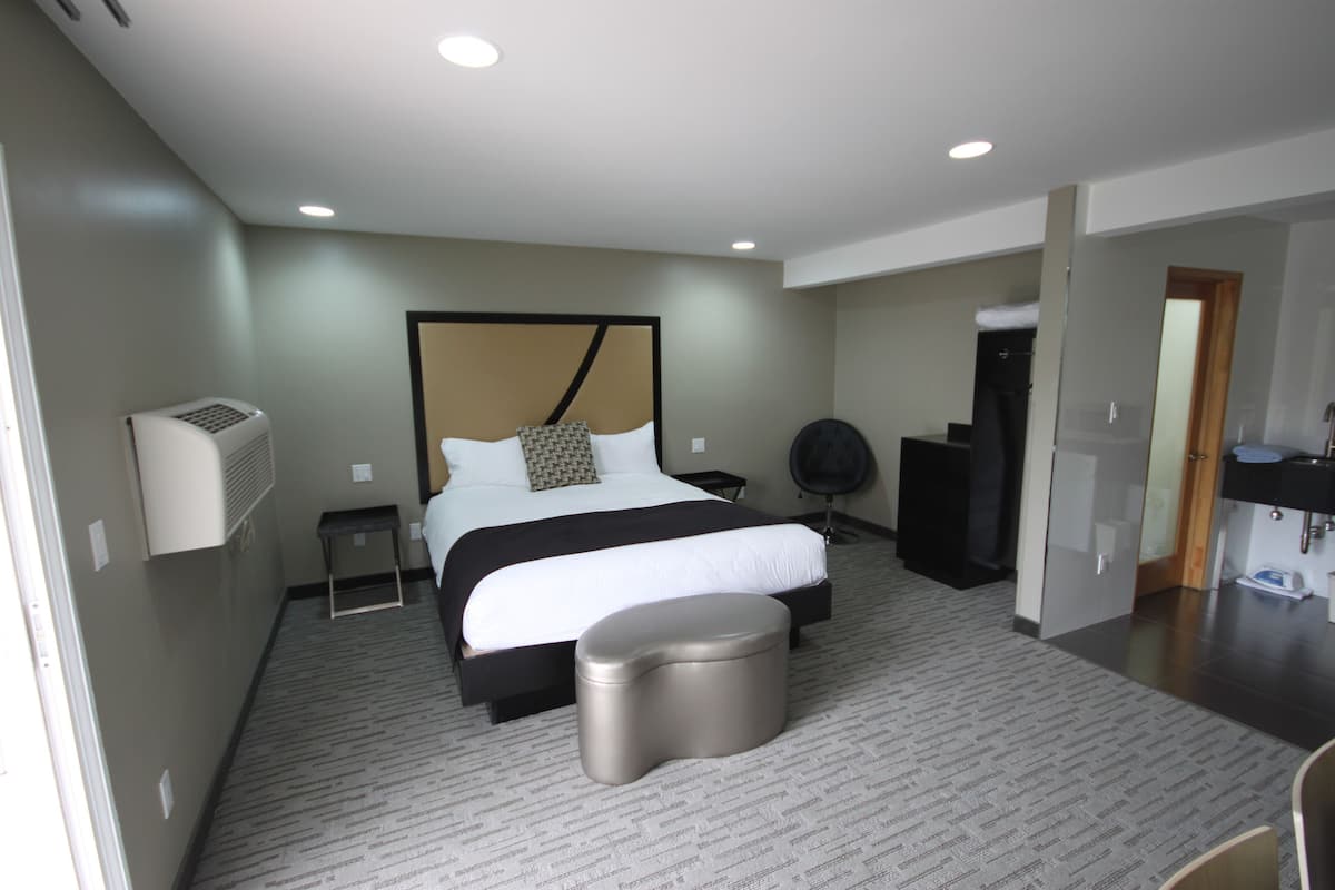 A modern hotel room with a large bed, a small bedside table, a chair, a trash bin, and a bathroom with a sink and mirror.