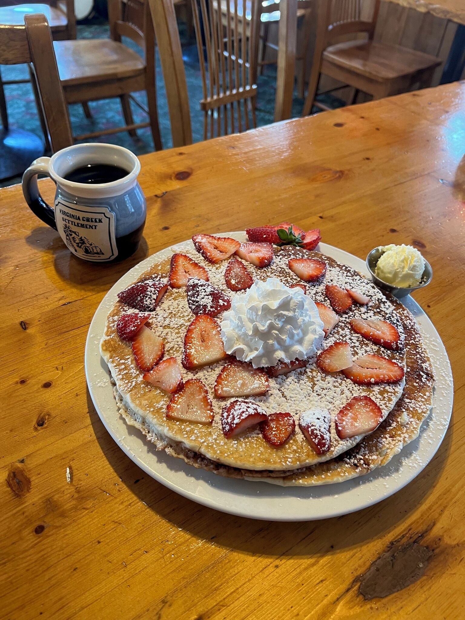 A plate of pancakes topped with strawberries, whipped cream, and powdered sugar, accompanied by a cup of coffee on a wooden table.
