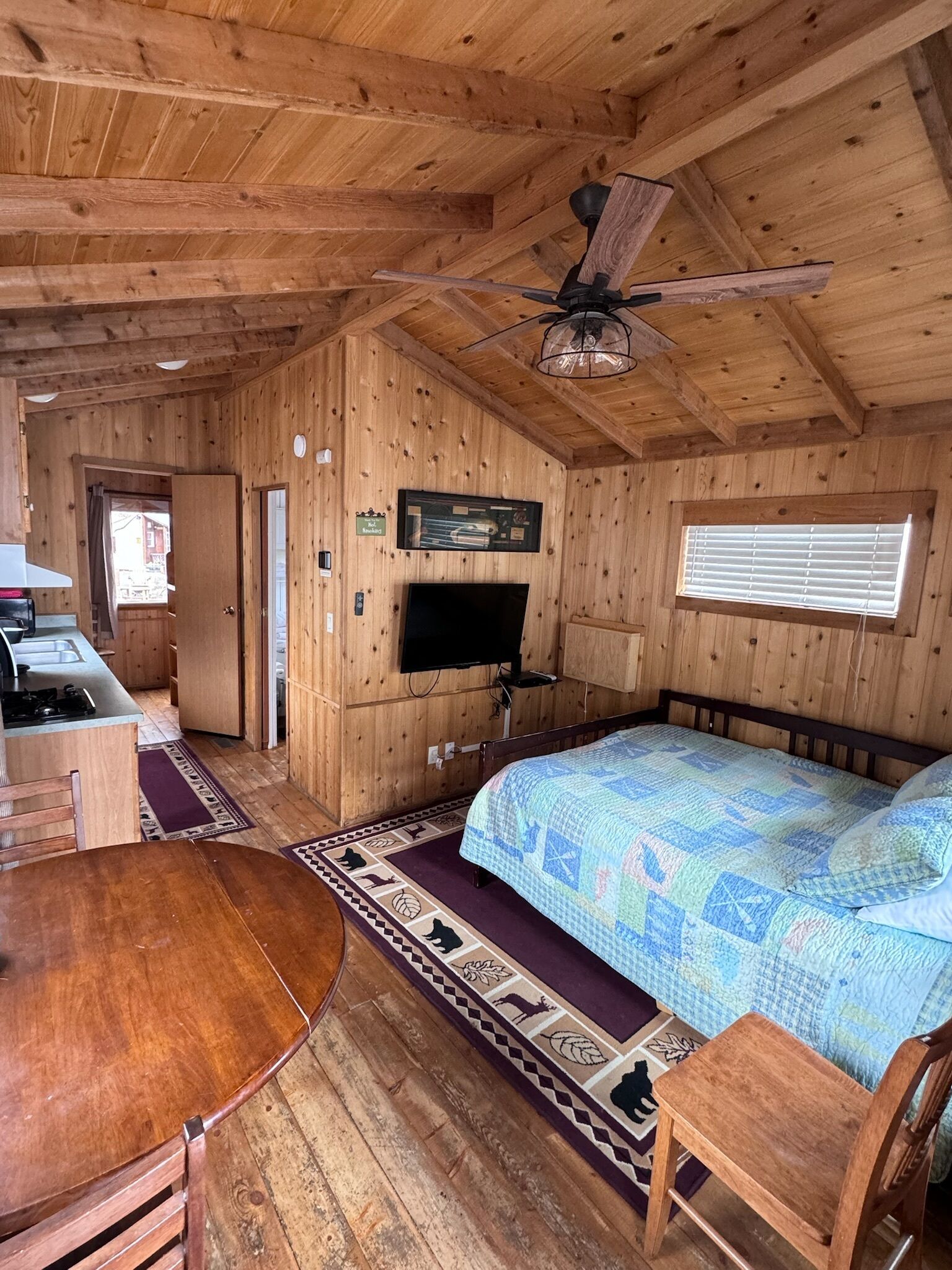 A wooden cabin with a bed, a TV, a ceiling fan, a dining table, and a kitchen area.