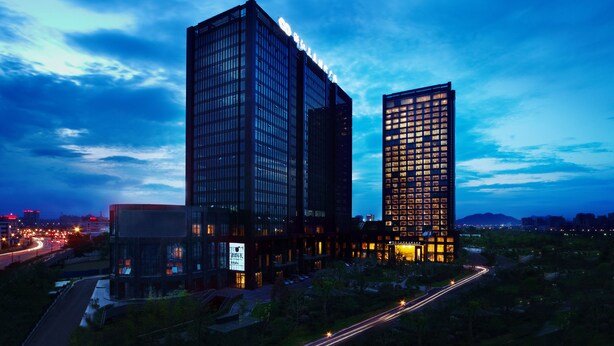 A modern high-rise building with a prominent glass facade and illuminated windows, surrounded by greenery and a busy road with light trails. - Grand New Century Hotel Canal Hangzhou (Hangzhou)