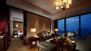 A spacious hotel room with a large bed, a sitting area with a sofa and armchairs, a dining table with a vase of flowers, and a view of the ocean through large windows. - Grand New Century Hotel Canal Hangzhou (Hangzhou)