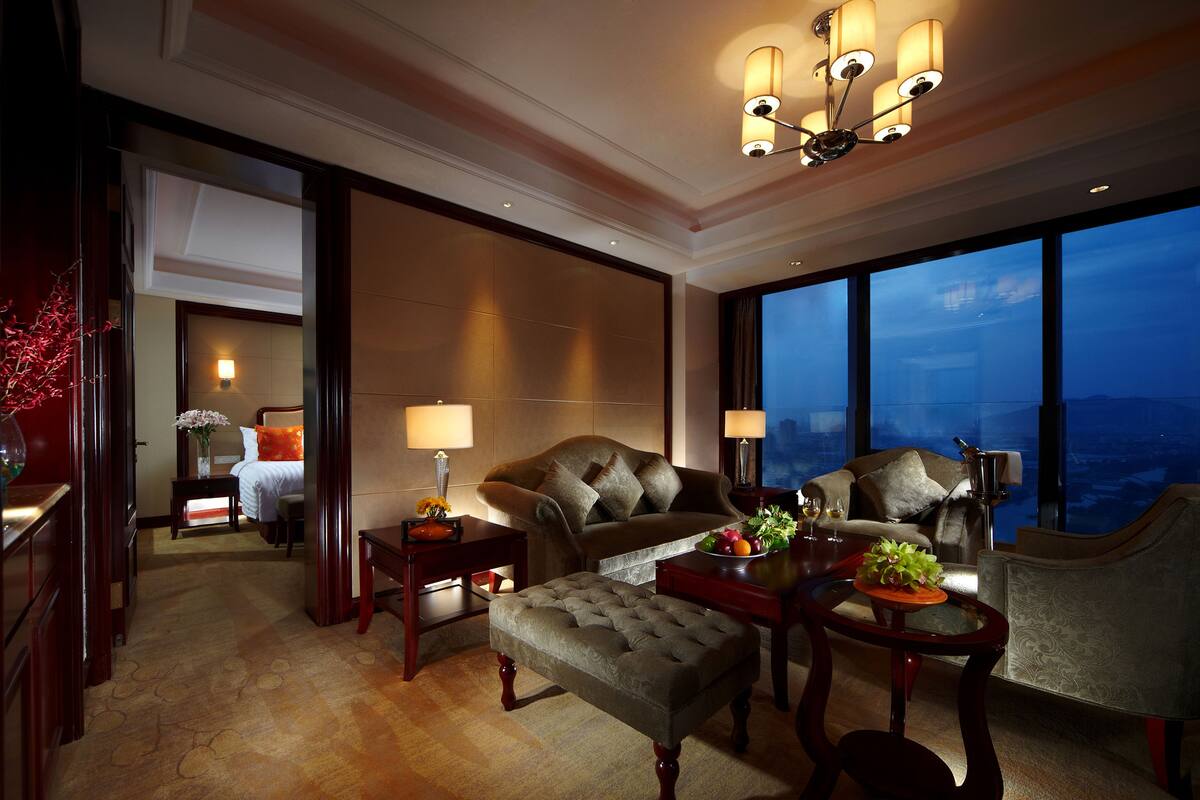Ein geräumiges Hotelzimmer mit einem großen Bett, einer Sitzecke mit einer Couch und Sesseln, einem Esstisch mit Blumenvase und Meerblick durch große Fenster.