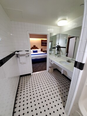 Une salle de bain d’hôtel avec un sol carrelé, un grand miroir, un lavabo et un espace douche.