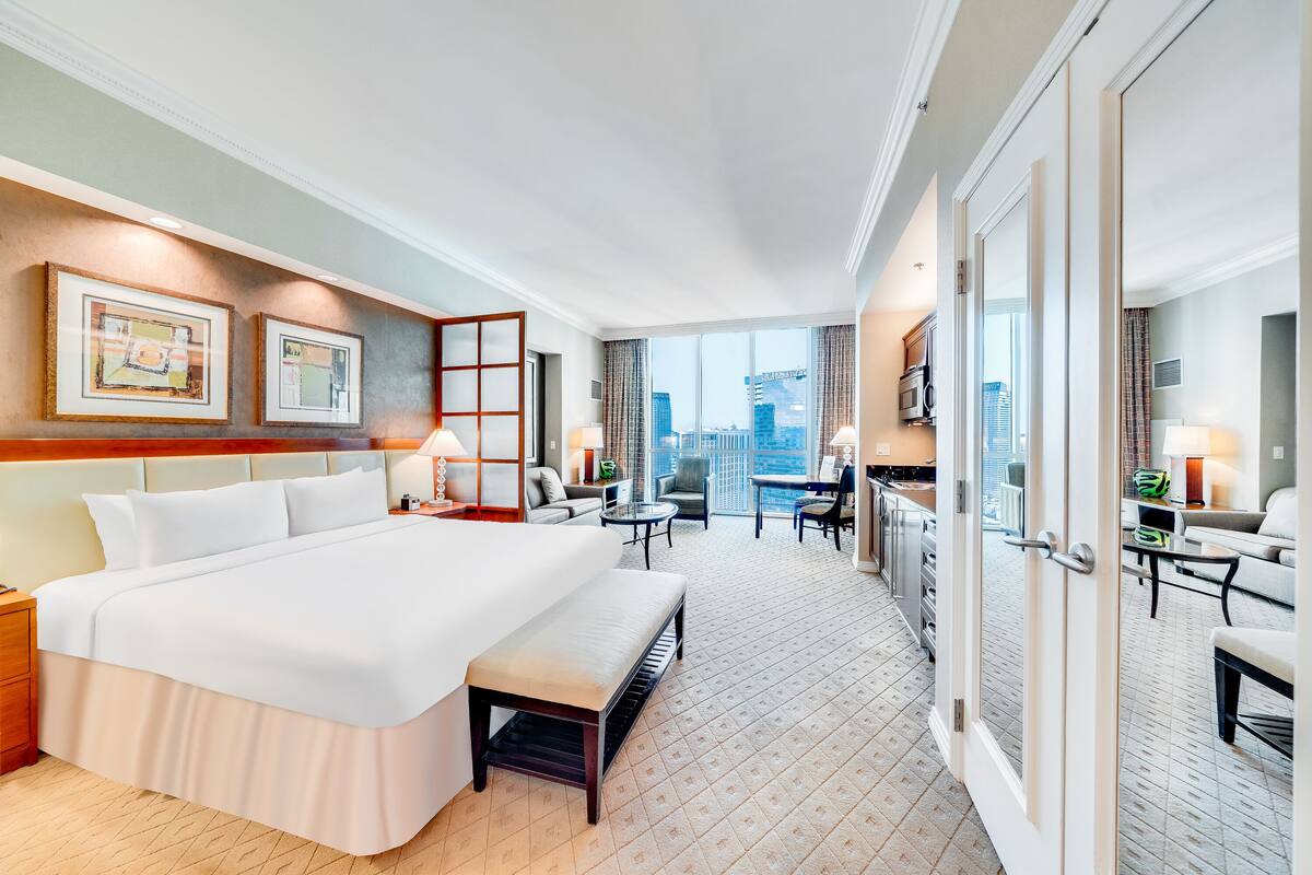 Habitación de hotel con una cama grande, un banco, un escritorio y vista a la ciudad.