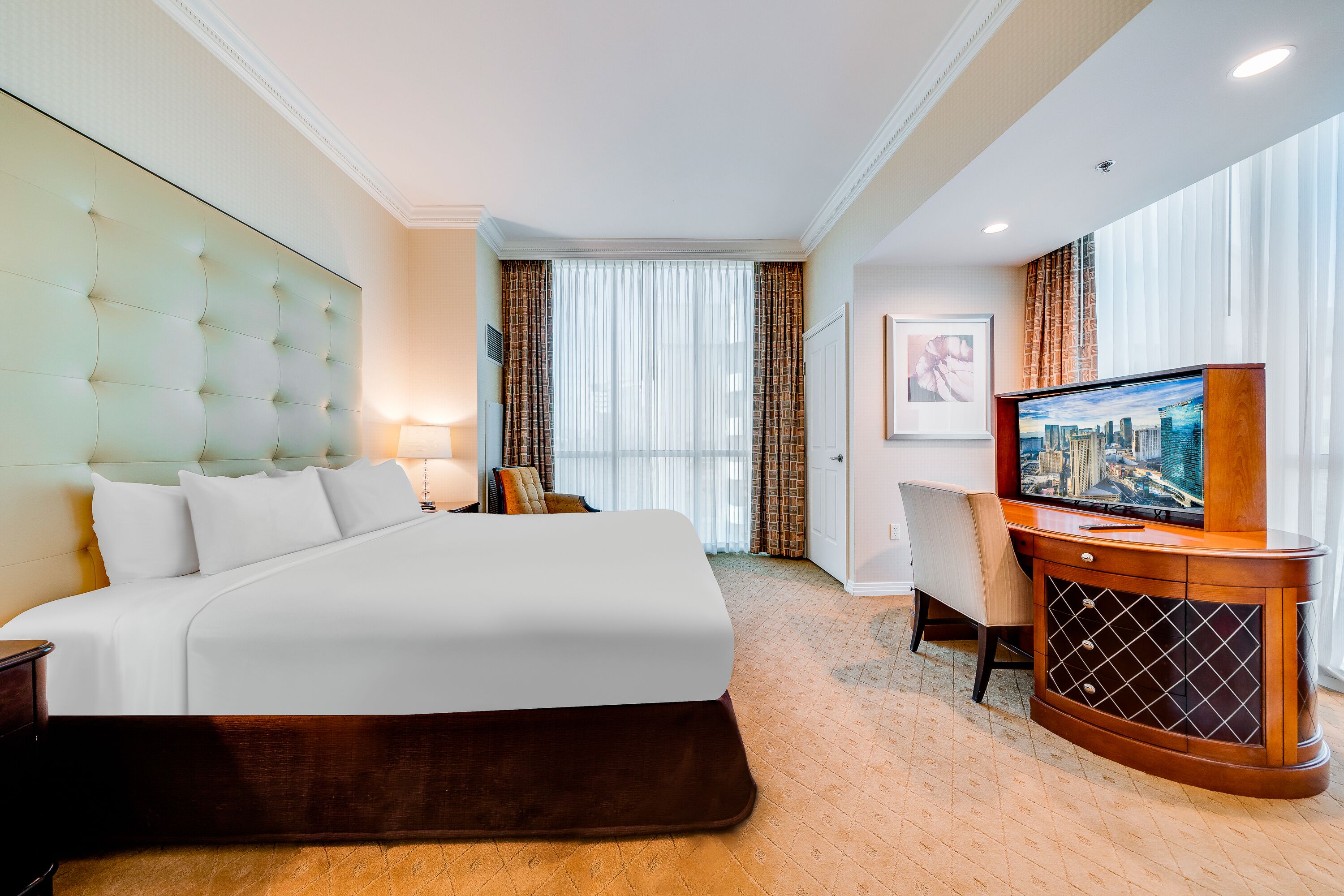 Ein Hotelzimmer mit einem großen Bett, einem Fernseher, einem Schreibtisch und einem Fenster mit Vorhängen.