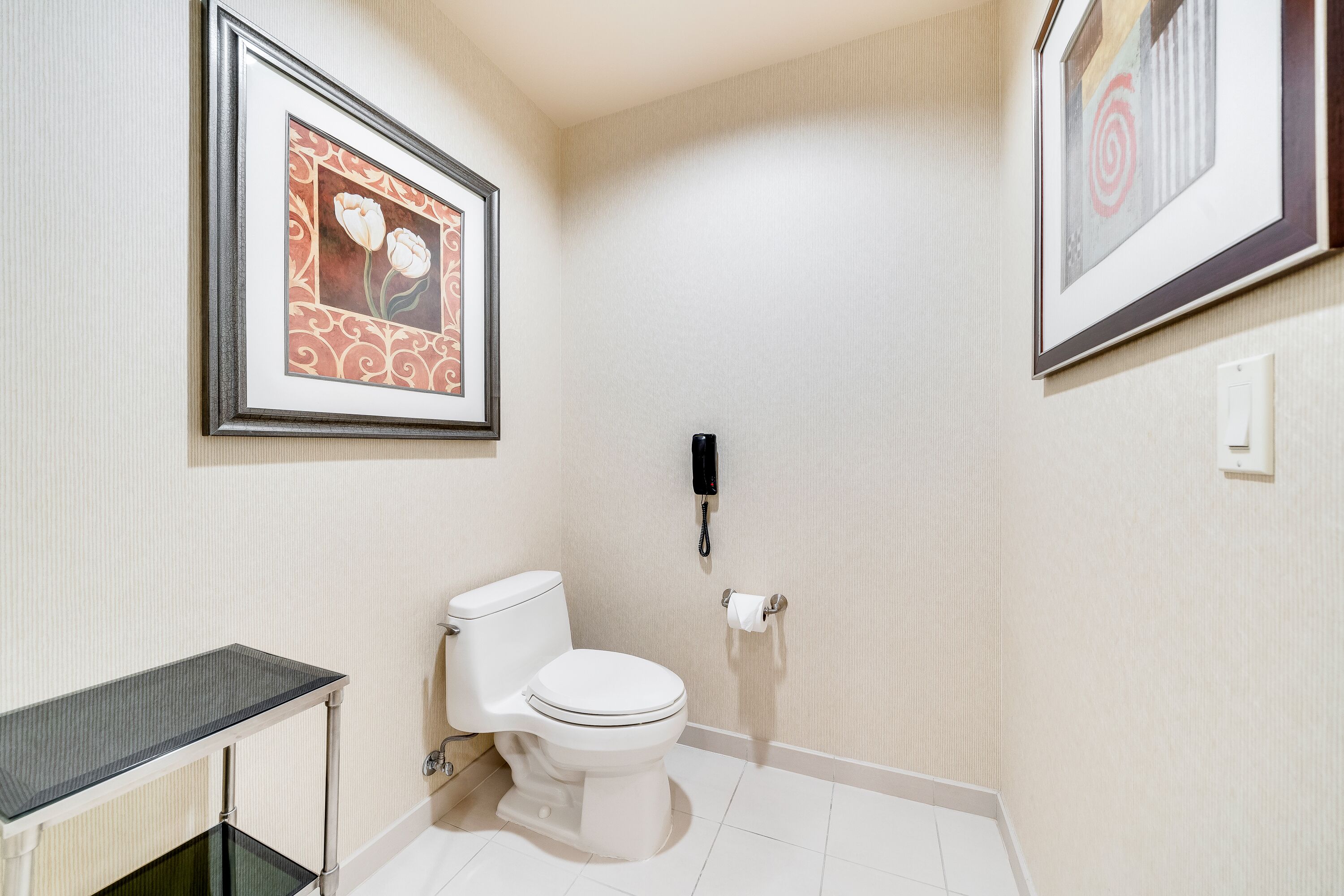 A bathroom with a toilet, a glass shelf, and two framed pictures on the wall.
