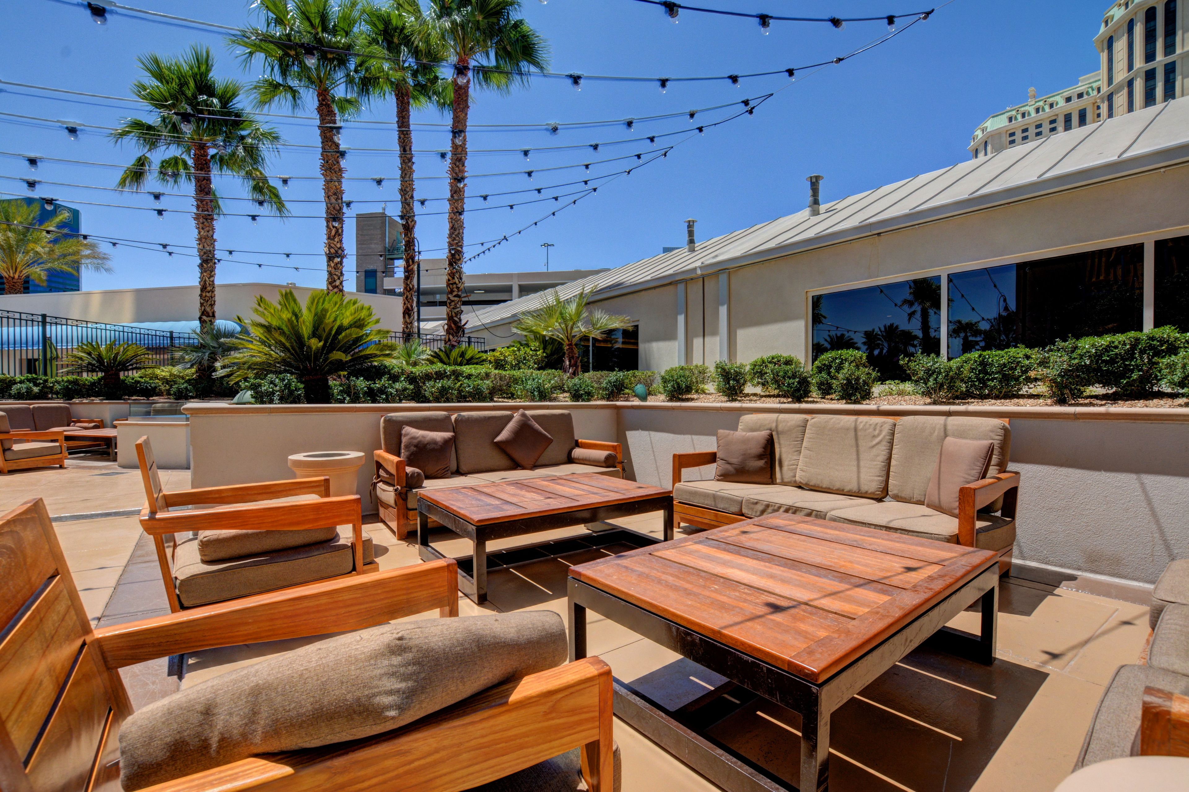 An outdoor patio area with wooden furniture, palm trees, and a building in the background.