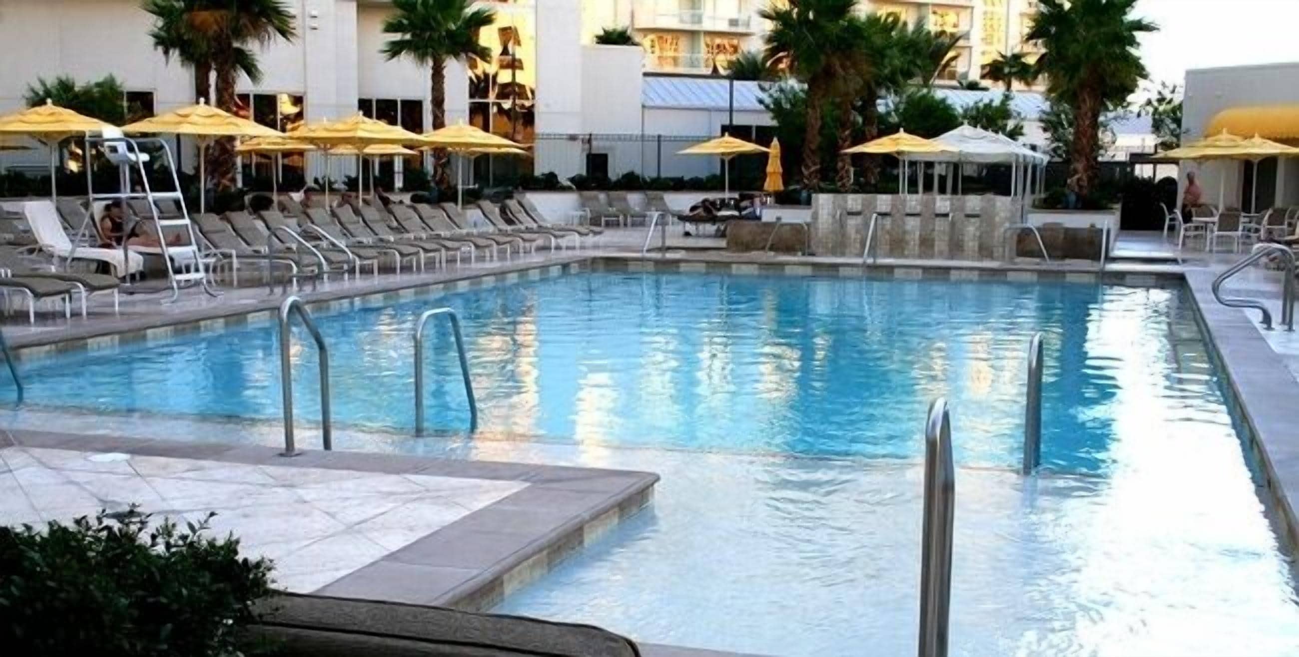 Ein Swimmingpool mit Liegestühlen und Sonnenschirmen, umgeben von Palmen und einem Gebäude im Hintergrund.