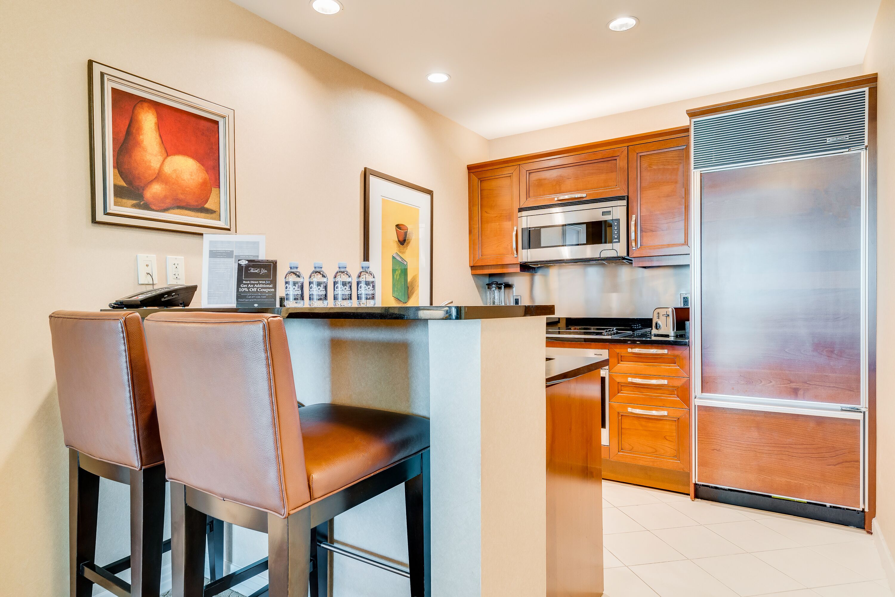 A modern kitchen with wooden cabinets, a stainless steel refrigerator, and a bar area with two leather chairs.