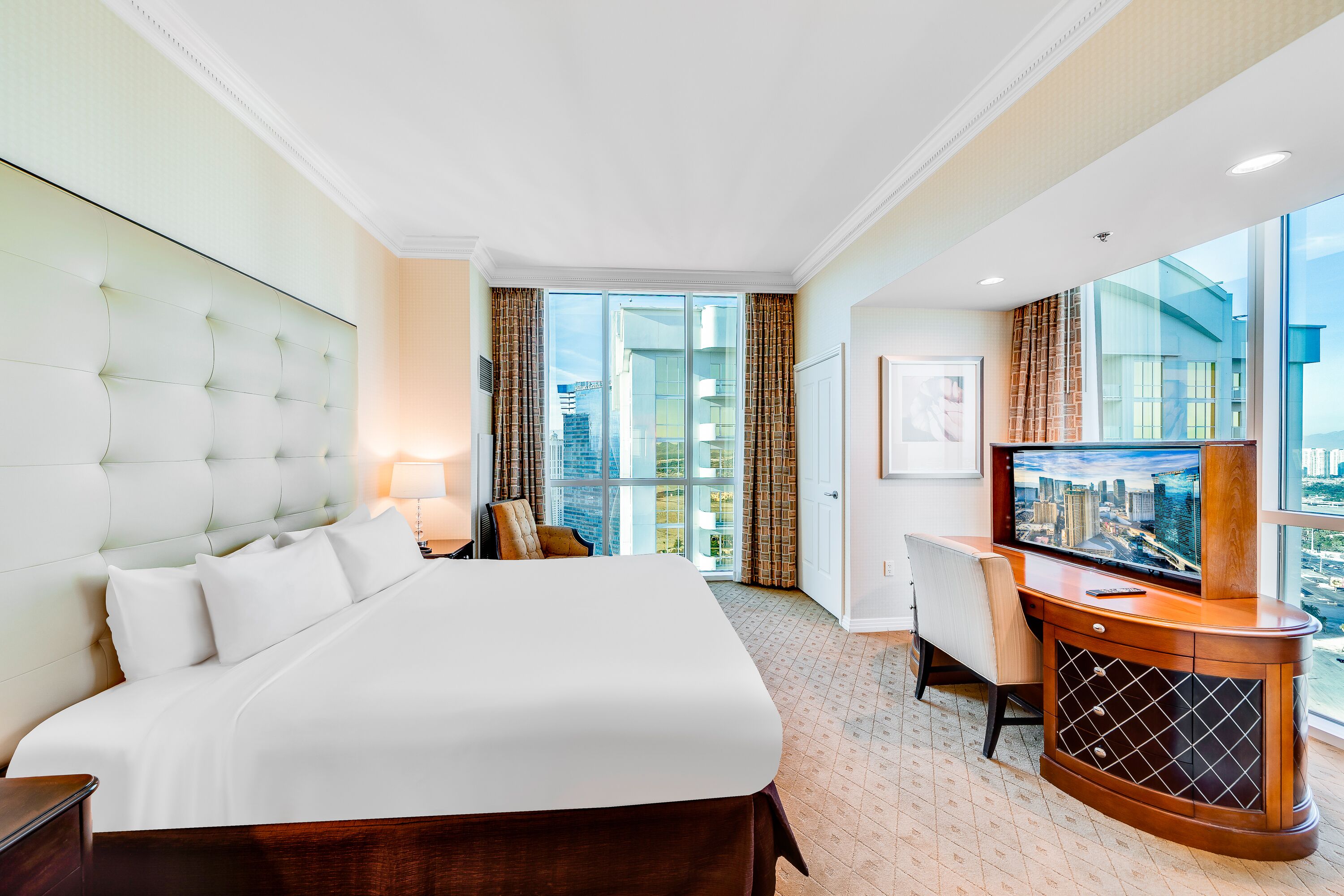 Een hotelkamer met een groot bed, een bureau met een televisie en uitzicht op de stad door grote ramen.