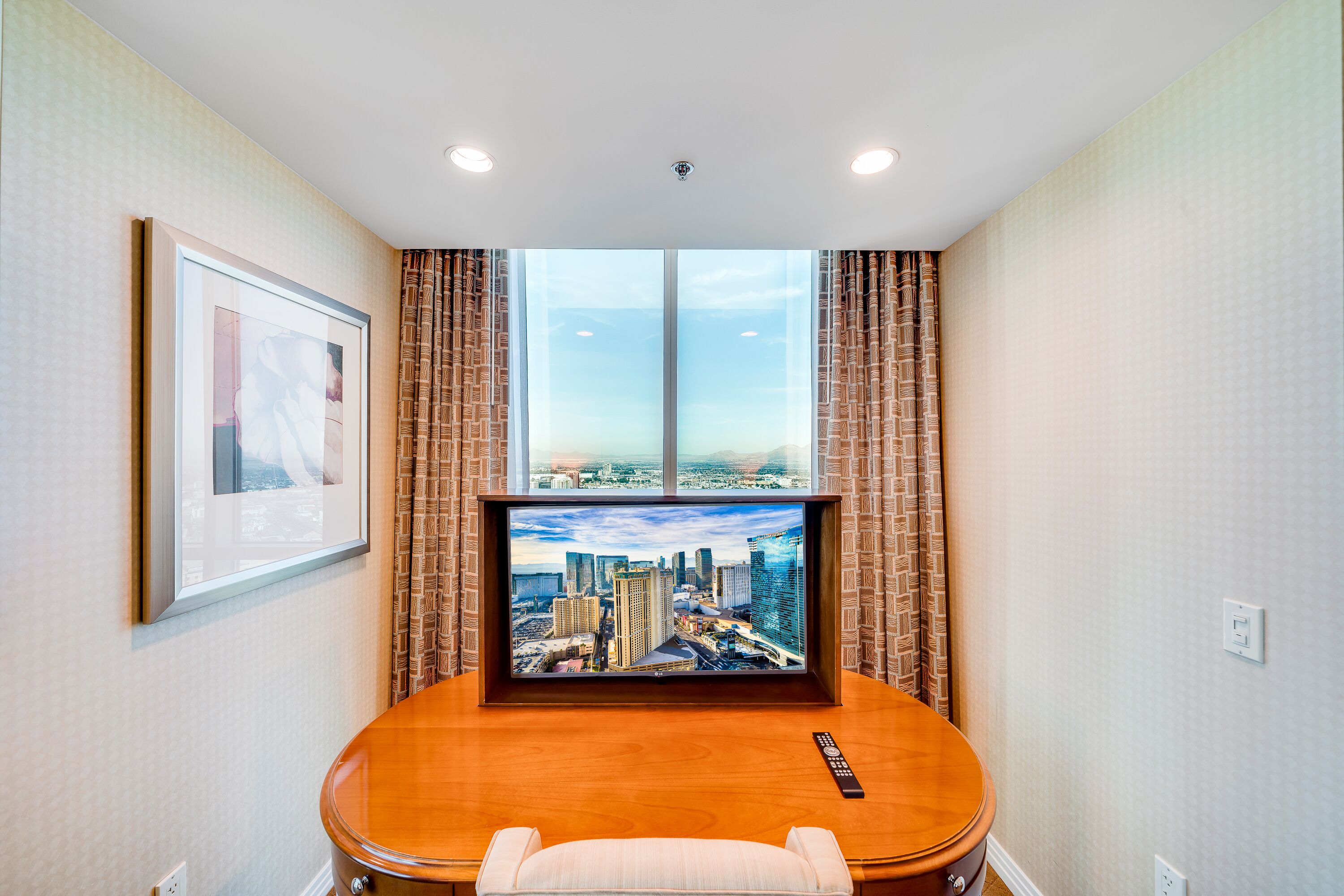 Une chambre d’hôtel avec une grande fenêtre, un tableau représentant un paysage urbain encadré, une table en bois et une télécommande.