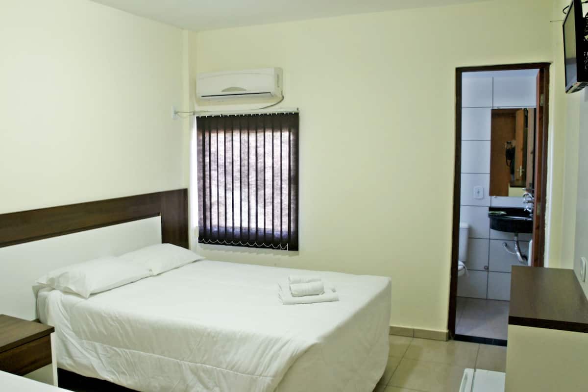 Ein Hotelzimmer mit Bett, Nachttisch, einem Fenster mit Jalousien, einer Klimaanlage und einem durchgegangenen Badezimmer.