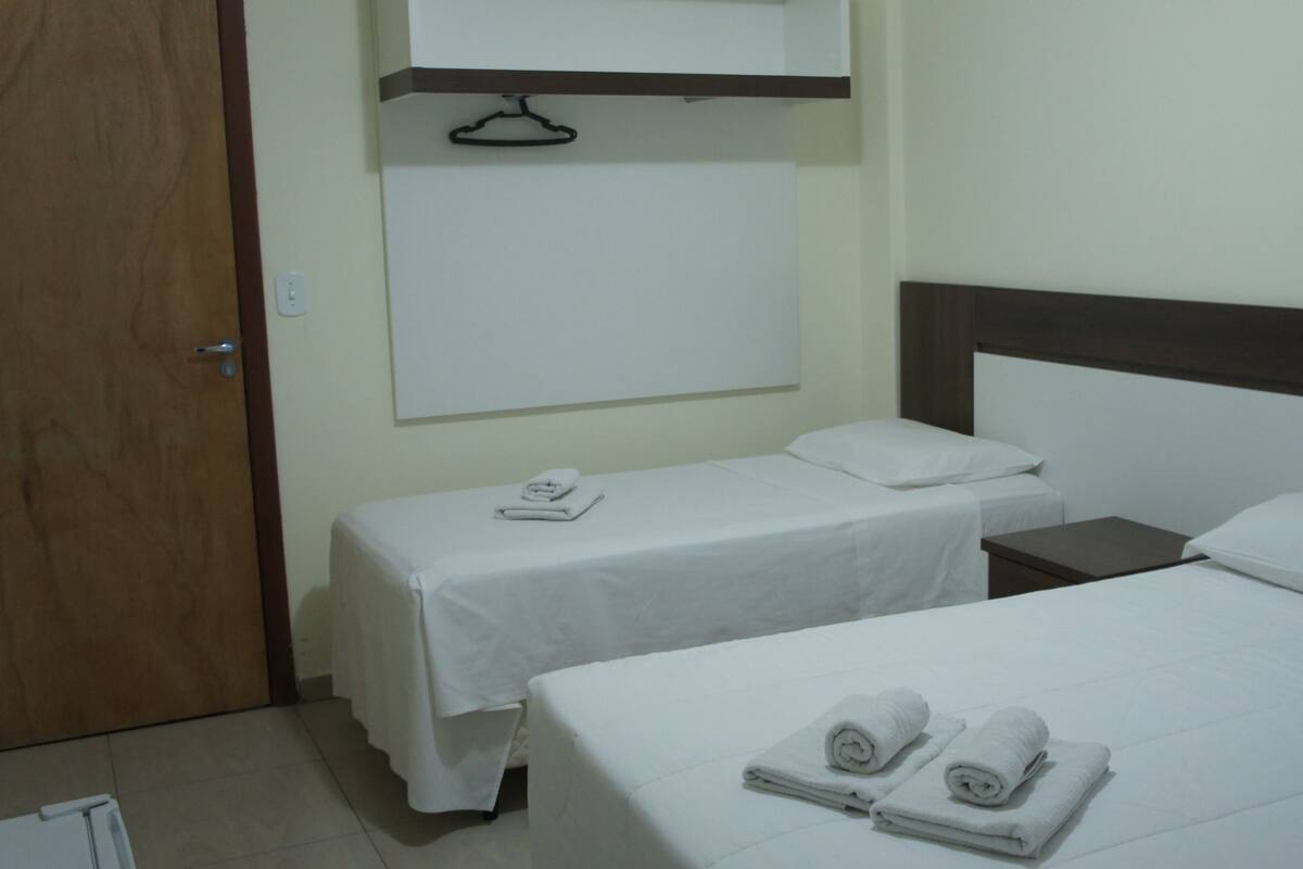 Ein Hotelzimmer mit zwei Einzelbetten, jeweils mit weißer Bettdecke und Kissen, und einem Holzkopfende. An der Wand über den Betten befindet sich eine Whiteboard und ein Kleiderständer.
