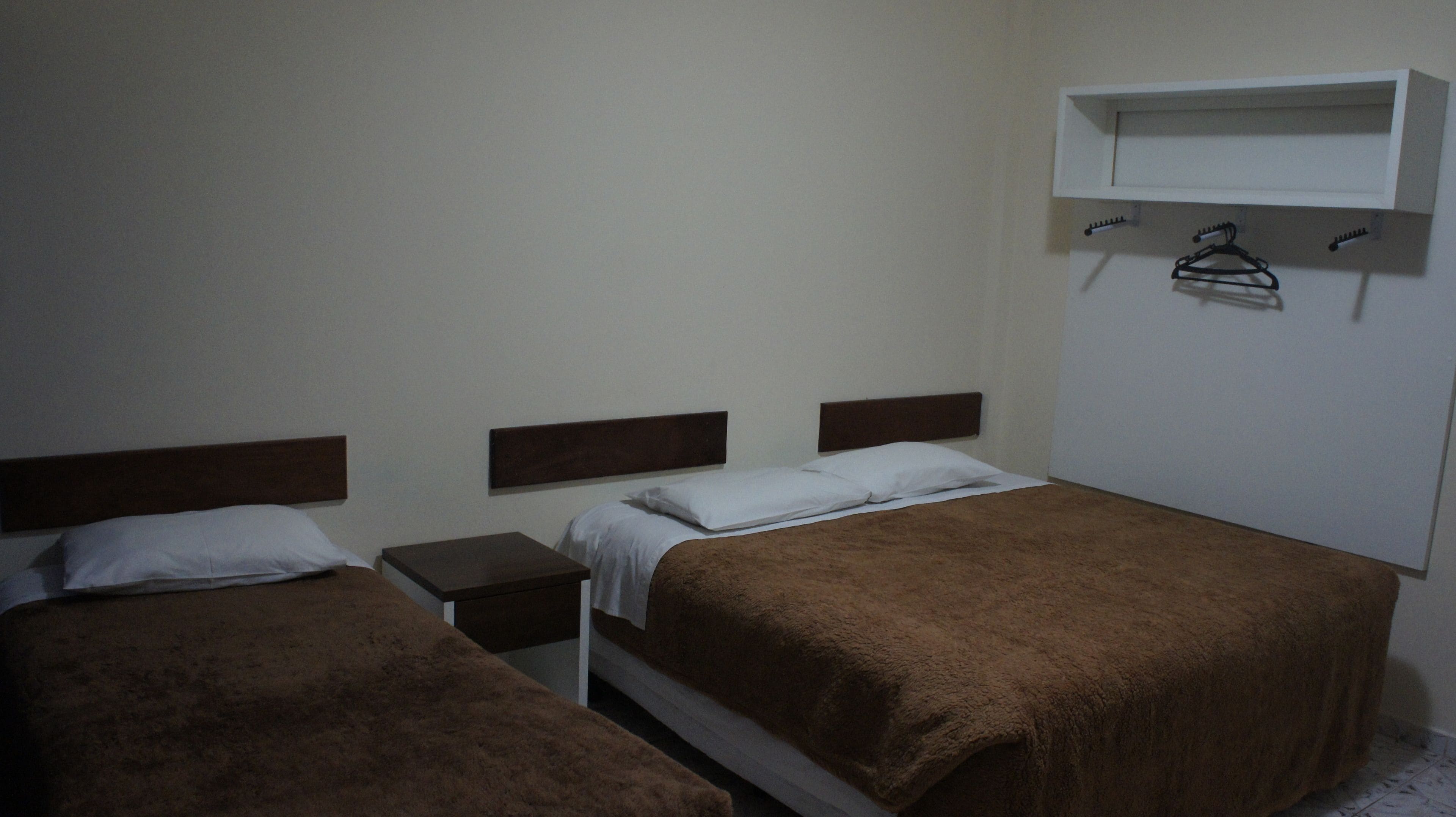 A simple room with two beds, each with a wooden headboard and a brown blanket. There is a small bedside table with a lamp on one side and a shelf above the bed on the other.