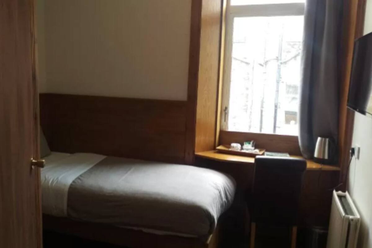 A hotel room with a bed, a nightstand, a window with a view of buildings, and a radiator.