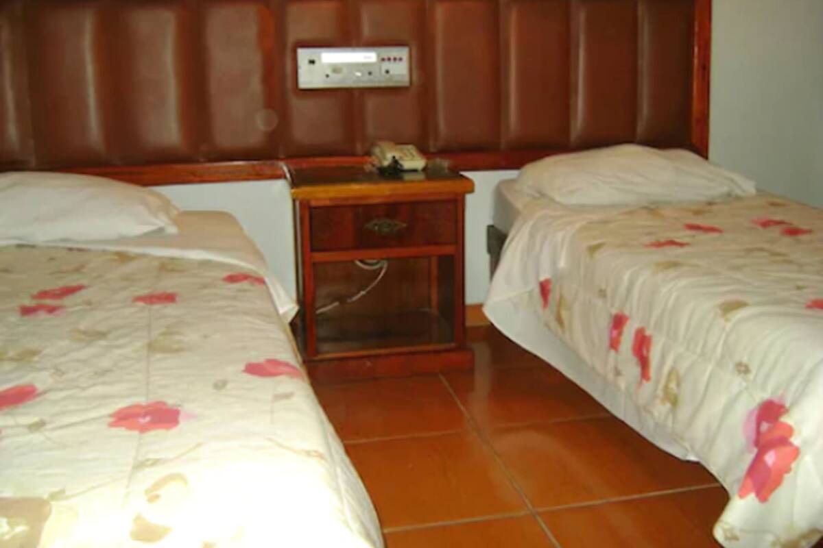 A hotel room with two single beds, a wooden headboard, a nightstand with a lamp, and a tiled floor.