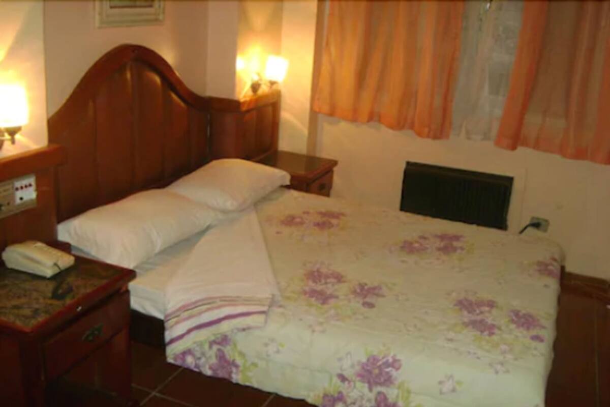 A hotel room with a wooden headboard, a floral bedspread, a bedside table with a lamp, and a nightstand with a phone.