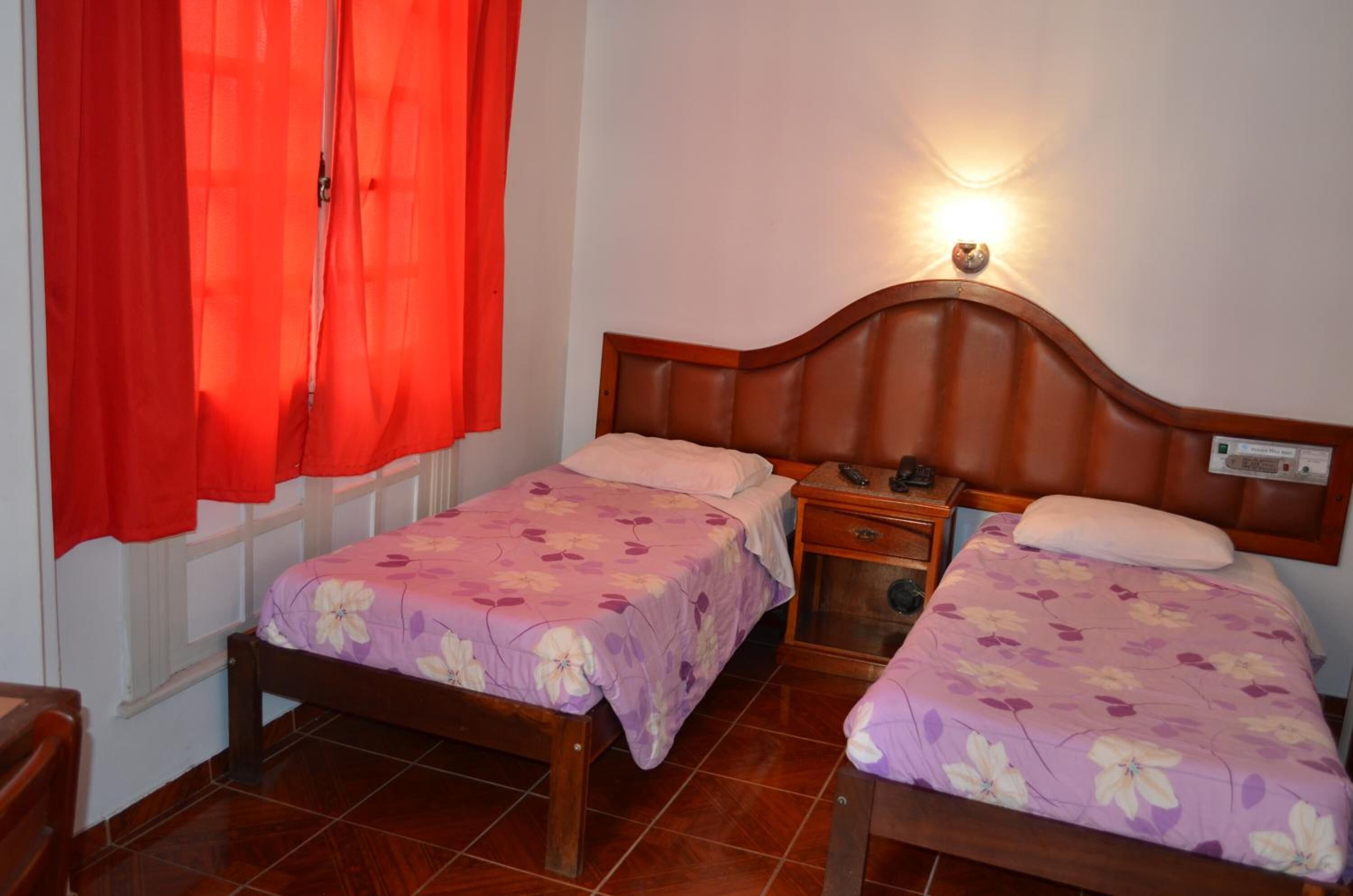 A room with two beds, each with a floral bedspread, a wooden headboard, a bedside table with a lamp, and a window with red curtains.