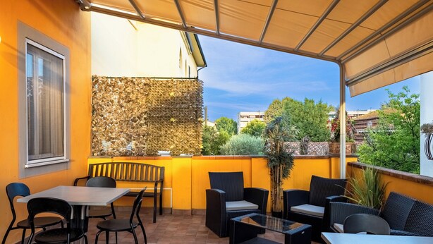 An outdoor patio area with a brick floor, yellow walls, and a variety of seating including tables and chairs. - B&B Alma (Lucca)