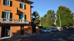 A two-story building with a storefront on the ground floor, featuring a sign that reads "B&B ALMA". There are two cars parked in front of the building, and a palm tree is visible to the right. - B&B Alma (Lucca)