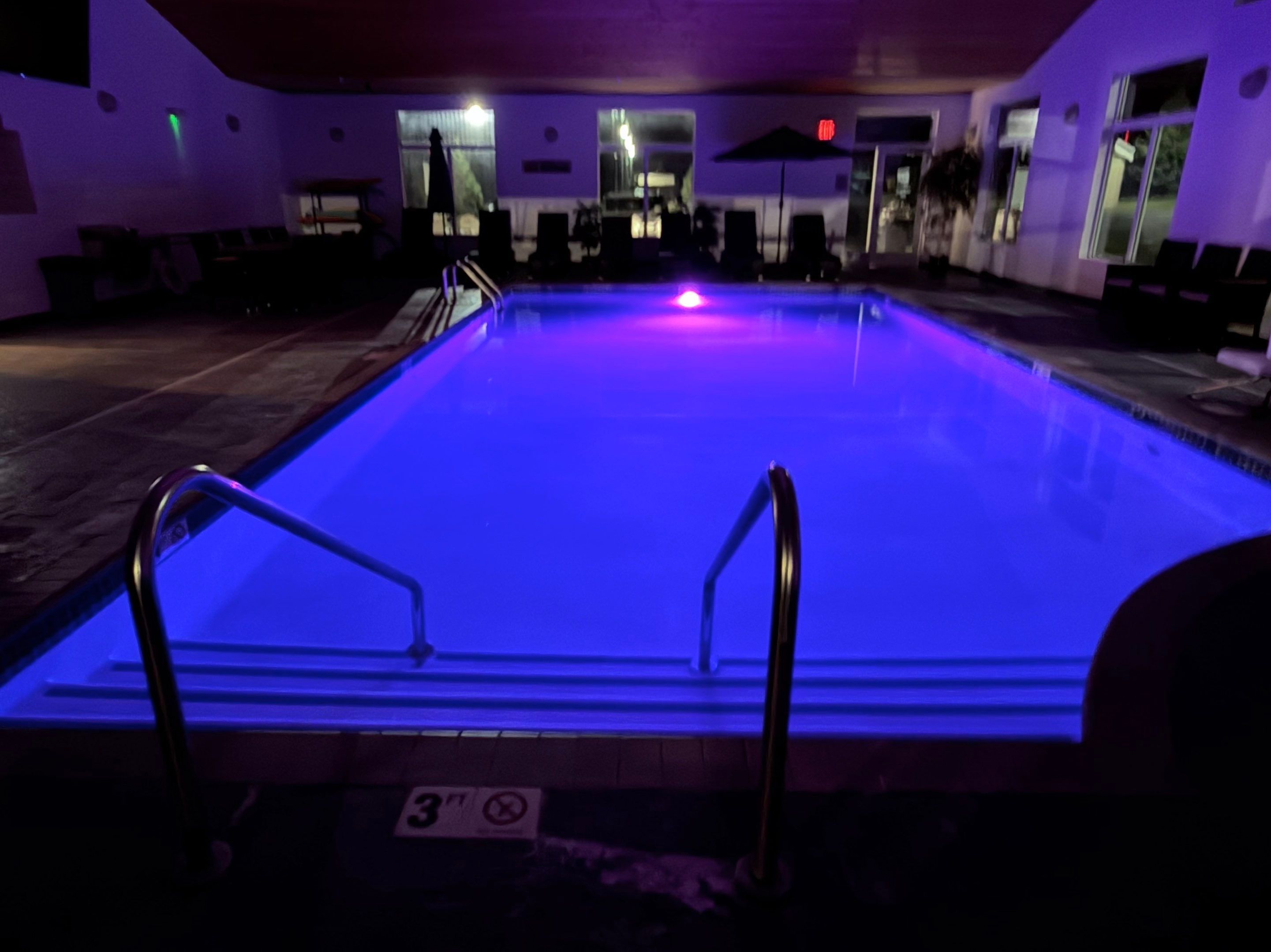 A swimming pool with a blue light, a 3ft no diving sign, and a lounge area with chairs and umbrellas.