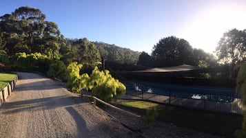 Ein Kiesweg führt zu einem überdachten Bereich mit einem Pool, umgeben von Bäumen und blauem Himmel.
